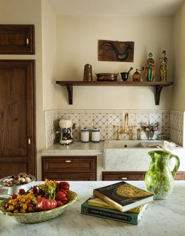 7 Best Agriturismos in Tuscany 16 A cozy kitchen with wooden cabinets, marble countertops, a farmhouse sink, and open shelves displaying pottery—evoking the warmth of agriturismos in Tuscany. On the counter are a green pitcher, cookbooks, and a bowl of colorful fruits and tomatoes.