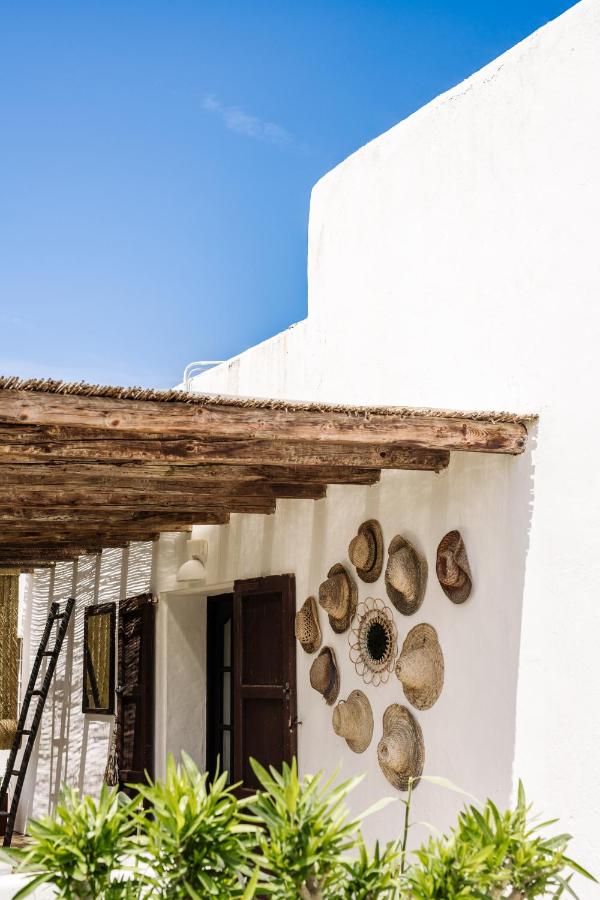 10 AUTHENTIC Finca Hotels in Ibiza 13 A white, Mediterranean-style building with a rustic wooden pergola, reminiscent of charming finca hotels in Ibiza. Straw hats are artfully arranged on the wall, and green plants line the foreground under a clear blue sky.