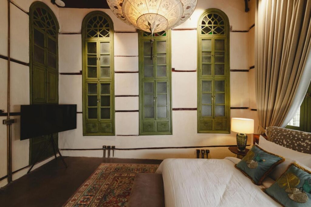 9 Best Hotels in Saudi Arabia: Unique Stays Across the Country 4 A cozy bedroom reminiscent of the Best Hotels in Saudi Arabia, featuring a white bed, patterned pillows, a decorative lamp, colorful rug, and four tall green-arched windows. A flat-screen TV and side table with a lamp complete the inviting space.