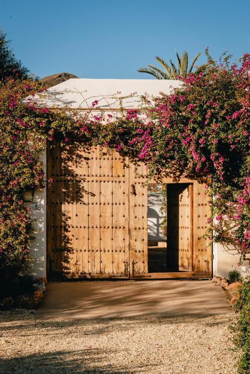 Adults-Only Hotels in Costa del Sol 8 A large wooden gate with a smaller door open within it is framed by vibrant pink and green flowering vines climbing up white walls, recalling the serene entrances of adult only hotels in Costa del Sol under a clear blue sky.
