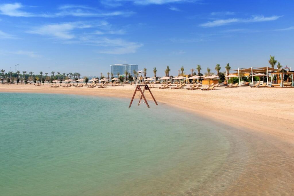 Where to Stay in Doha, Qatar: Transit, City & Beach Hotels 17 A sandy beach with clear turquoise water, sun loungers, and umbrellas—perfect for relaxing while you explore where to stay in Doha. Palm trees and a few buildings are visible in the background under a bright blue sky with wispy clouds.