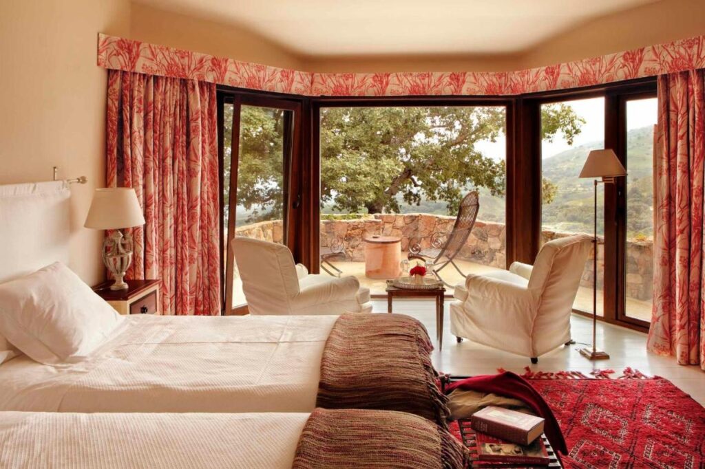 25 Weekend Trips from Madrid Max. 2h Away 38 A cozy bedroom with two single beds, armchairs by large windows, red-patterned curtains, and a rug—perfect for weekend trips from Madrid. Outside, a stone patio with table and chairs offers a scenic view of trees and hills.