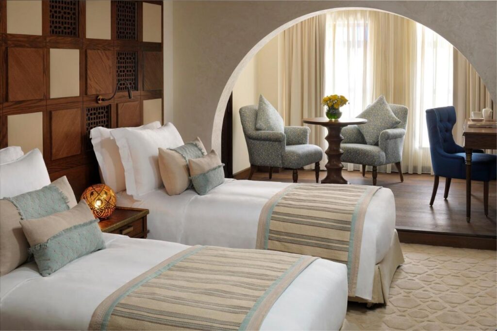 Where to Stay in Doha, Qatar: Transit, City & Beach Hotels 36 A cozy hotel room—perfect for those seeking where to stay in Doha—with two single beds, decorative pillows, a wooden accent wall, and a seating area featuring armchairs, a table with a yellow flower vase, and a desk by large curtained windows.