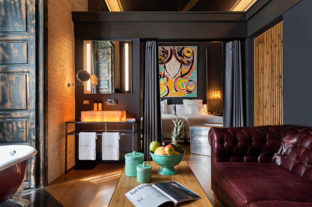 10 UNIQUE Hotels with Pool in Seville 3 A stylish hotel suite—with a burgundy leather sofa, freestanding bathtub, vanity with orange sink, fruit bowl on a wooden table, and a bed beneath colorful abstract art—offers refined comfort among the best hotels with pool in Seville.