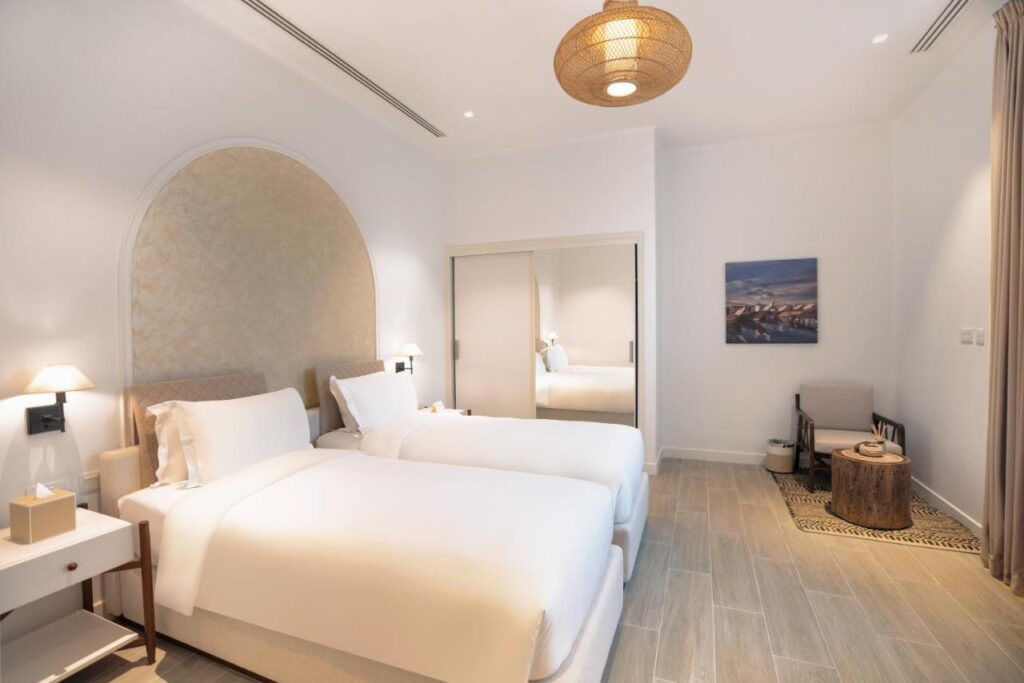 Where to Stay in Doha, Qatar: Transit, City & Beach Hotels 35 A bright, modern hotel room—perfect for those searching where to stay in Doha—features two single beds with white bedding, a woven ceiling lamp, mirror-fronted closet, small table, armchair, and minimalist neutral decor.