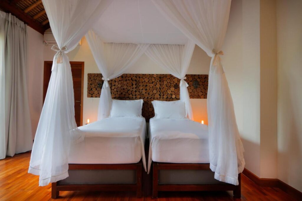 Two twin beds with white bedding and a shared mosquito net canopy are set side by side in a bright room with wooden floors—a perfect setup you'll find in some of the best family villas in Bali.