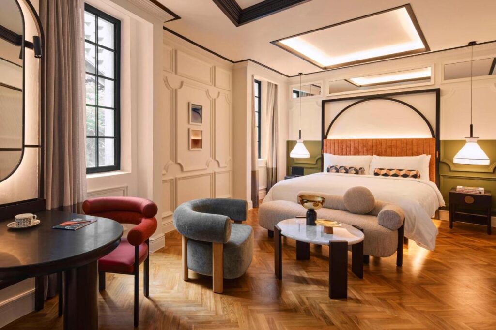 13 Most Unique Hotels in Singapore for an Incredible Holiday 28 Modern hotel room at one of the unique hotels in Singapore with a large bed, stylish armchairs, small tables, parquet floor, and tall windows. Neutral tones with green and wood accents plus soft lighting create a cozy atmosphere.