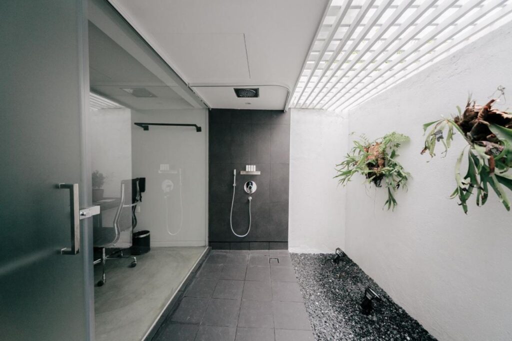 13 Most Unique Hotels in Singapore for an Incredible Holiday 14 Modern open-air shower in one of the unique hotels in Singapore, featuring a dark tiled wall, glass door entrance, minimalist fixtures, and potted plants on a white wall under a slatted roof that lets in natural light.