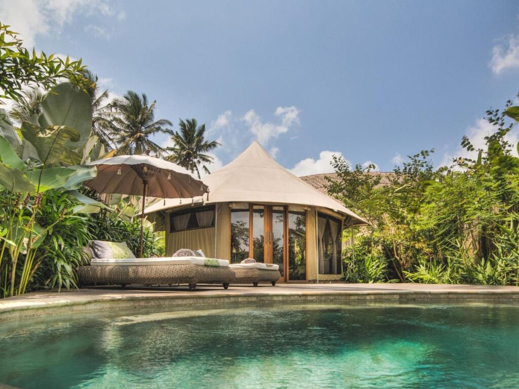 13 Most UNIQUE Places to Stay in Ubud 30 A luxurious villa with a pointed canvas roof sits behind a turquoise pool, surrounded by lush tropical plants, palm trees, and a sun lounger with an umbrella—one of the most unique places to stay in Ubud under a bright blue sky.