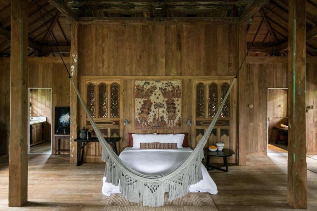 13 Most UNIQUE Places to Stay in Ubud 6 A cozy wooden bedroom with a bed and decorative pillows, a bohemian-style hammock in front, carved wooden panels, and a large tapestry above the headboard—one of the unique places to stay in Ubud. Two doorways lead to other rooms.