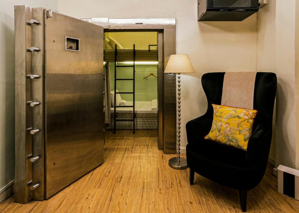 13 Most Unique Hotels in Singapore for an Incredible Holiday 45 A cozy room with a large open vault door, a black armchair with a yellow floral pillow, and a floor lamp. A glimpse of a bunk bed and ladder hints at creative design—perfect for fans of unique hotels in Singapore. The floor is wooden.