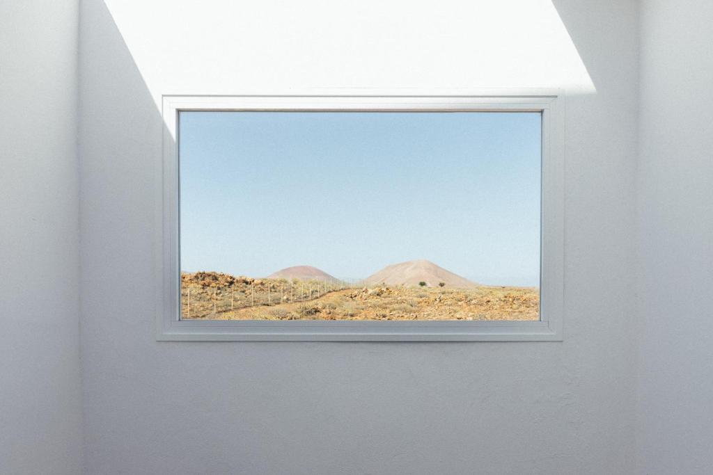Unique Adults Only Hotels in Lanzarote 21 A bright, minimalist white room in one of the Adults Only Hotels in Lanzarote features a large window framing distant hills under a clear blue sky and a dry, rocky landscape. Sunlight casts geometric shadows on the pristine walls.