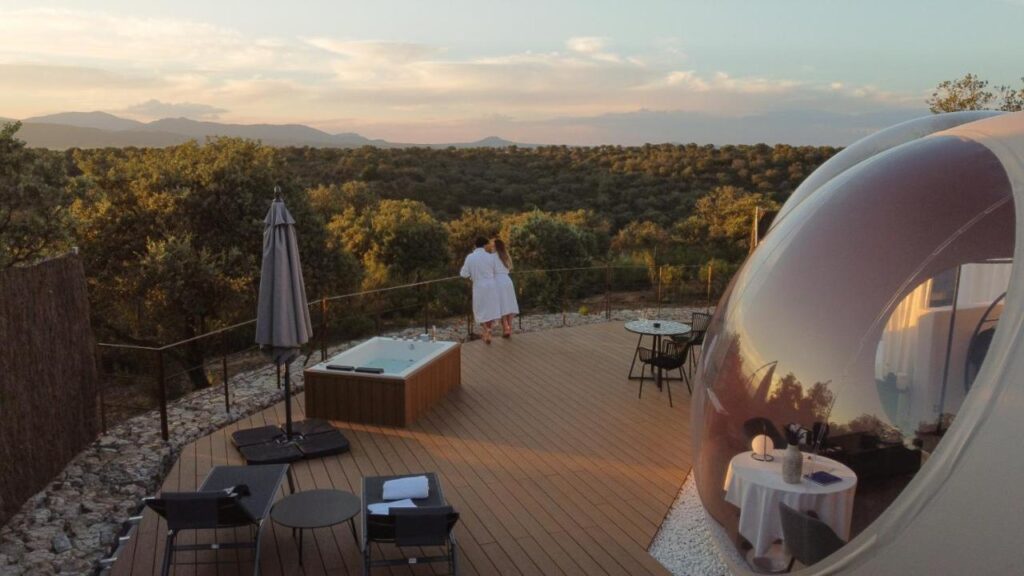 25 Weekend Trips from Madrid Max. 2h Away 18 A person in a white robe stands on a wooden terrace with outdoor seating, a hot tub, and a bubble-shaped transparent dome—an ideal spot for weekend trips from Madrid—overlooking a forested landscape at sunset.