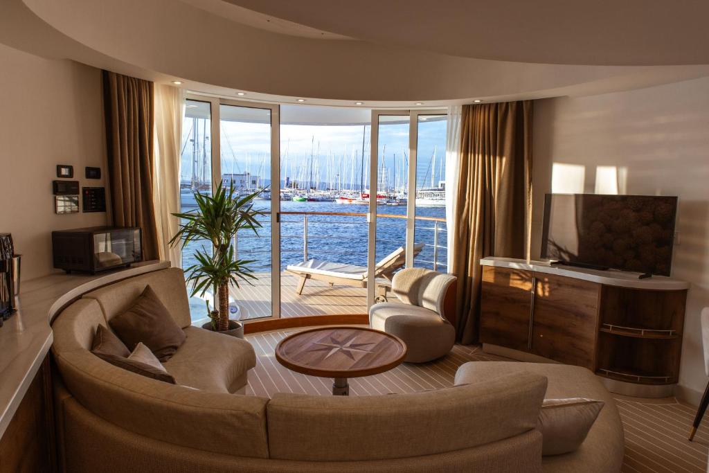 Where to Stay in Split: Hotel & Areas Guide 35 A modern, curved living room with beige furniture, a TV, and large windows opening to a balcony overlooking a marina with docked sailboats—an ideal spot if you’re wondering where to stay in Split. Natural light brightens the cozy, elegant space.