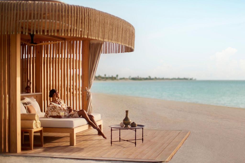 9 Best Hotels in Saudi Arabia: Unique Stays Across the Country 31 A woman relaxes on a lounge chair under a wooden cabana by a sandy beach at one of the Best Hotels in Saudi Arabia. A small table and decorative vases sit nearby, while the calm sea and clear sky create a tranquil, tropical setting.