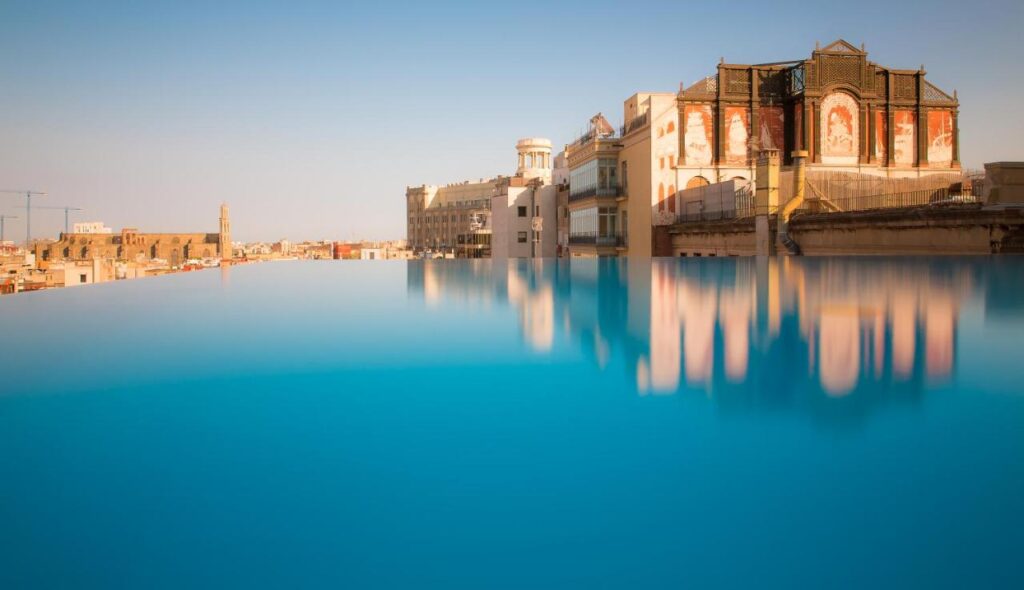 Top Barcelona Hotels with Rooftop Pool 25 A rooftop infinity pool at one of the finest Barcelona Hotels with Rooftop Pool overlooks a cityscape of historic buildings and clear blue sky, reflecting the city's unique architecture and sunlight on its calm water surface.