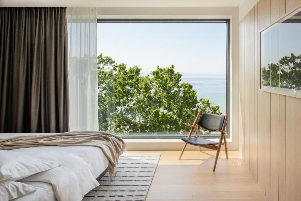 8 TOP Boutique Hotels in Split 26 A modern bedroom, perfect for those researching where to stay in Split, with a large window overlooking green trees and water. The space features white bedding, a beige throw, striped rug, wooden chair, and light wood-paneled walls.