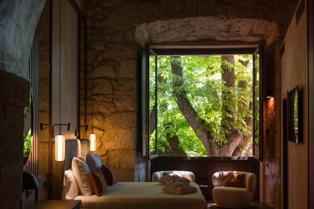 25 Weekend Trips from Madrid Max. 2h Away 96 A cozy bedroom with stone walls and soft lighting features a large open window revealing lush green trees outside—a perfect retreat for weekend trips from Madrid. The bed is neatly made, with a chair by the window creating a peaceful, inviting atmosphere.