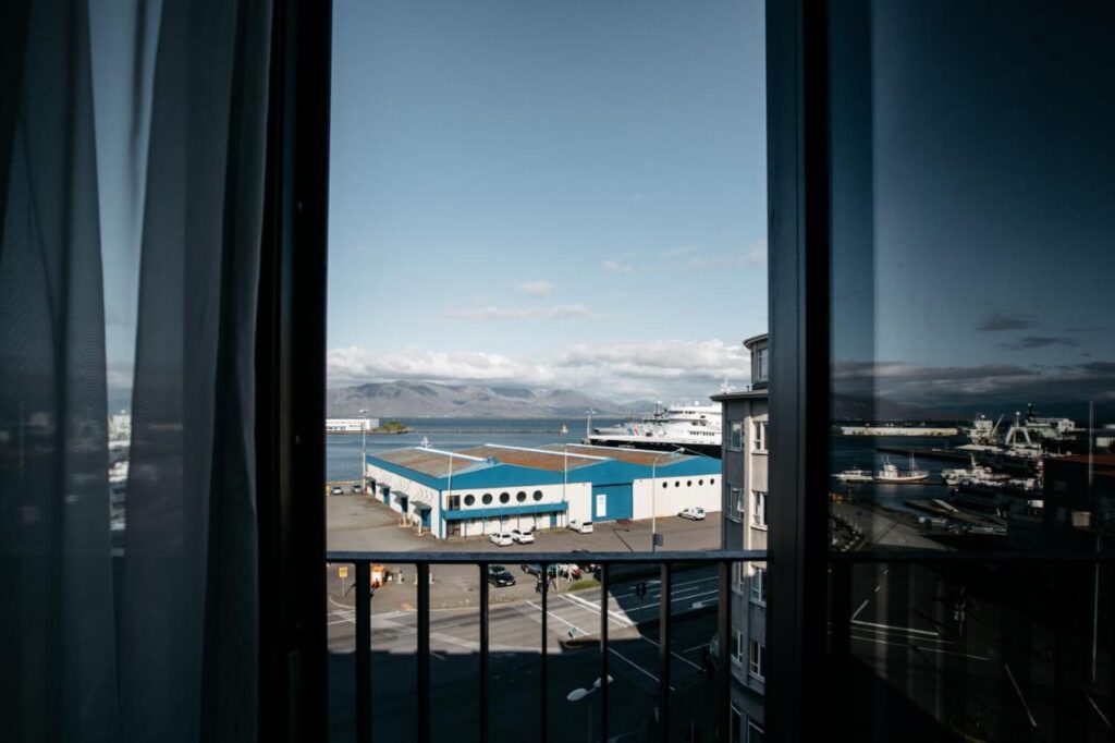 Where to Stay in Reykjavík: Top Guide 11 View through a window with sheer curtains, showing a blue and white industrial warehouse near a harbor in Reykjavik—an ideal scene for those wondering where to stay in Reykjavik, with mountains, partly cloudy sky, street, and parked cars below.