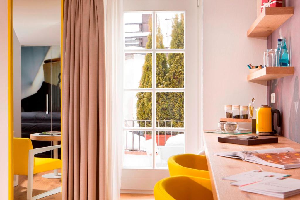Unique Boutique Hotels in Salzburg 31 A bright, modern room in the style of boutique hotels in Salzburg, featuring yellow chairs, a desk with books and a yellow kettle, shelves with glassware, and a large window with beige curtains overlooking trees outside.