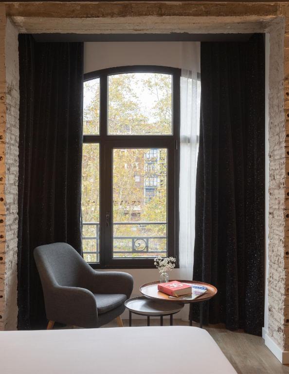 8 Unique Boutique Hotels in Bilbao Spain 7 A cozy reading nook, reminiscent of the inviting spaces found in boutique hotels in Bilbao, features a gray armchair and two small round tables by a large window framed with black curtains, offering views of autumn trees. A book and a small vase of flowers rest on the tables.