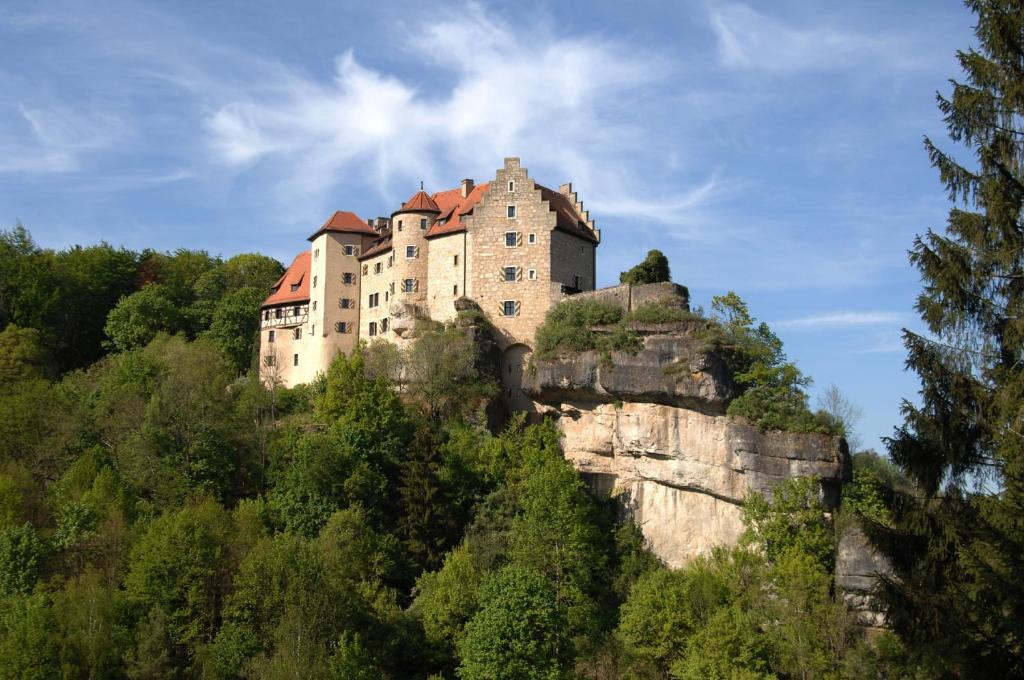 FAIRYTALE Castle Hotels in Bavaria 11 A stone castle with red-roofed towers, like the enchanting castle hotels in Bavaria, sits atop a rocky cliff, surrounded by dense green trees and set against a blue sky with wispy clouds.