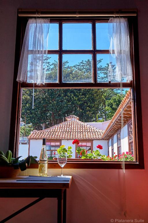 11 UNIQUE Boutique Hotels in Tenerife 25 A window with sheer white curtains opens to a view of red-roofed buildings and blooming red flowers—reminiscent of the charm found in Boutique Hotels in Tenerife. On the sill, a bottle of white wine and glass bask in the bright daylight streaming in.