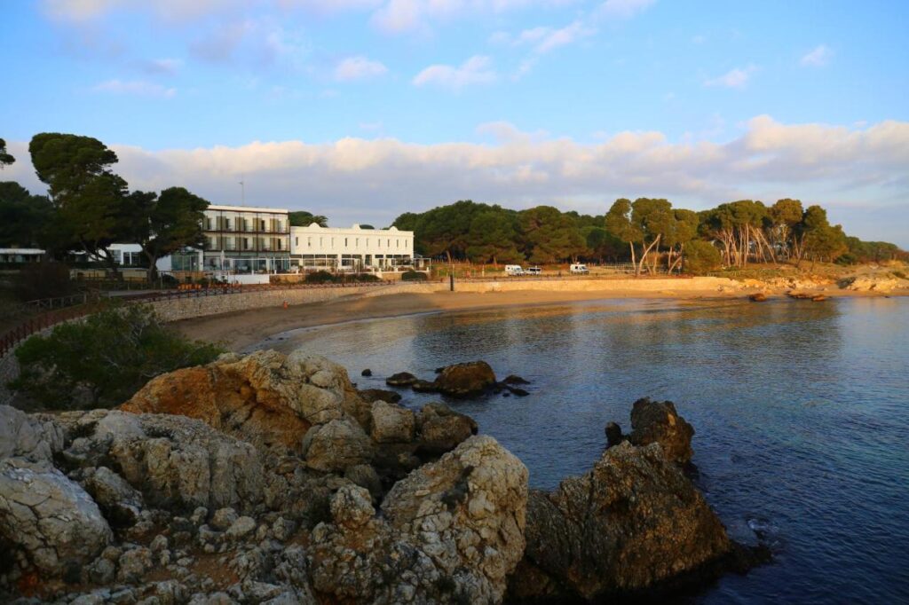 A scenic coastal view with a rocky foreground, calm sandy beach, and a white hotel building—one of the boutique hotels in Costa Brava—surrounded by trees under a partly cloudy sky at sunset.