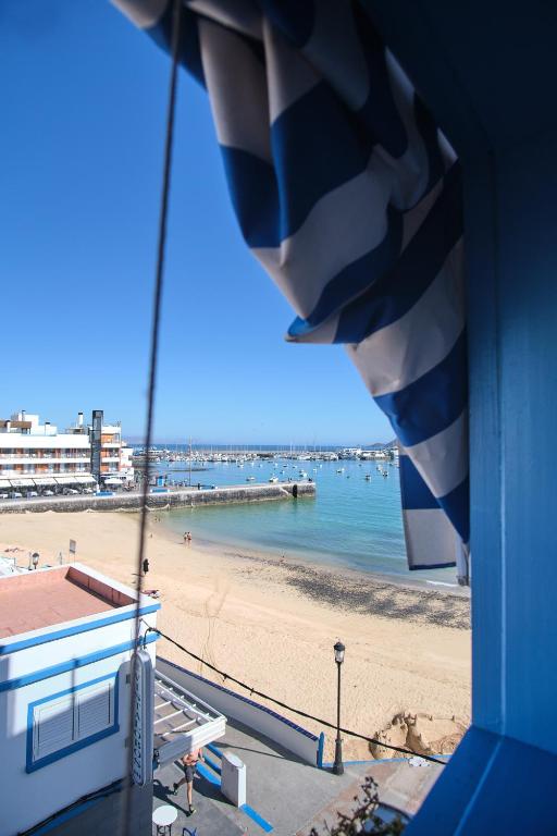 Adults Only Hotels in Fuerteventura 24 View of a sandy beach and calm blue sea from a window with a blue-and-white striped awning above. A few people stroll on the beach near adults only hotels in Fuerteventura, with buildings, a pier, and boats in the background.