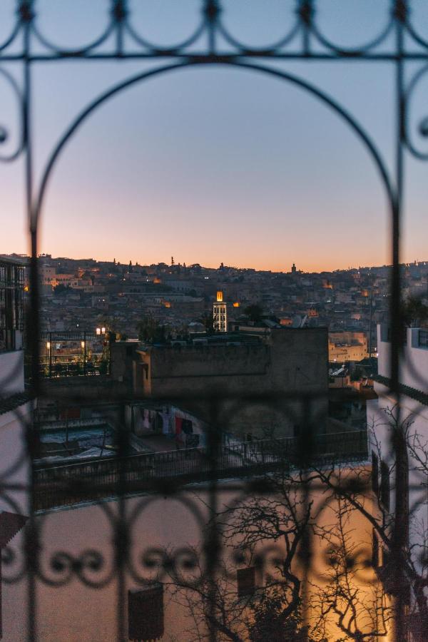 7 UNIQUE Riads in Fez You Won't Forget 21 View of a cityscape at sunset seen through a decorative wrought iron window, reminiscent of traditional riads in Fez. The orange and blue sky glows above distant buildings with a lit tower, while shadows and rooftops fill the foreground.