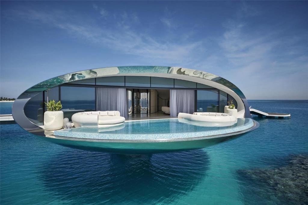 9 Best Hotels in Saudi Arabia: Unique Stays Across the Country 37 A futuristic, oval-shaped villa with floor-to-ceiling windows and a curved infinity pool floats above clear blue ocean water under a sunny sky—an experience reminiscent of the Best Hotels in Saudi Arabia. Stylish white outdoor furniture decorates the terrace.