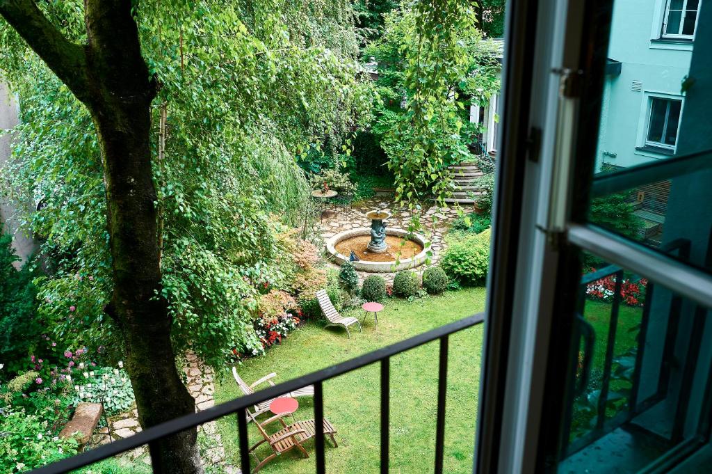 Unique Boutique Hotels in Salzburg 29 View from an open window at one of the finest Boutique Hotels in Salzburg, overlooking a lush garden with a circular stone fountain, green lawn, lawn chairs, small red tables, and vibrant flowers surrounded by trees.