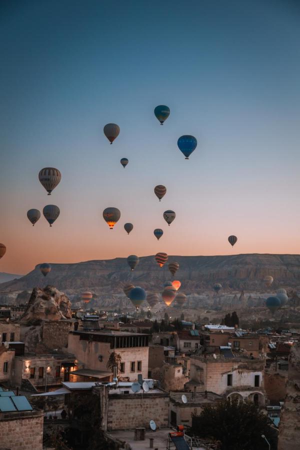 Most Authentic Cave Hotels in Cappadocia 4 Dozens of colorful hot air balloons float above a town with stone buildings and cave hotels in Cappadocia at sunrise, with rocky hills and a clear sky in the background.
