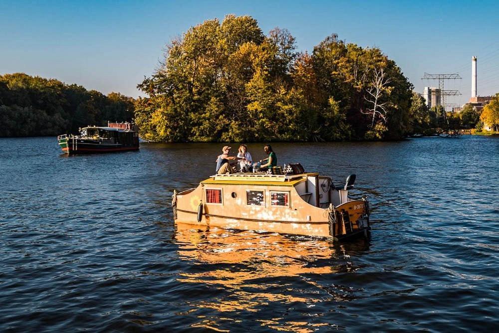 Boutique Hotels in Berlin Kreuzberg 40 A small houseboat with three people on its roof floats on a calm river, evoking the laid-back charm of boutique hotels in Berlin Kreuzberg, with trees and another boat in the background under a clear blue sky.
