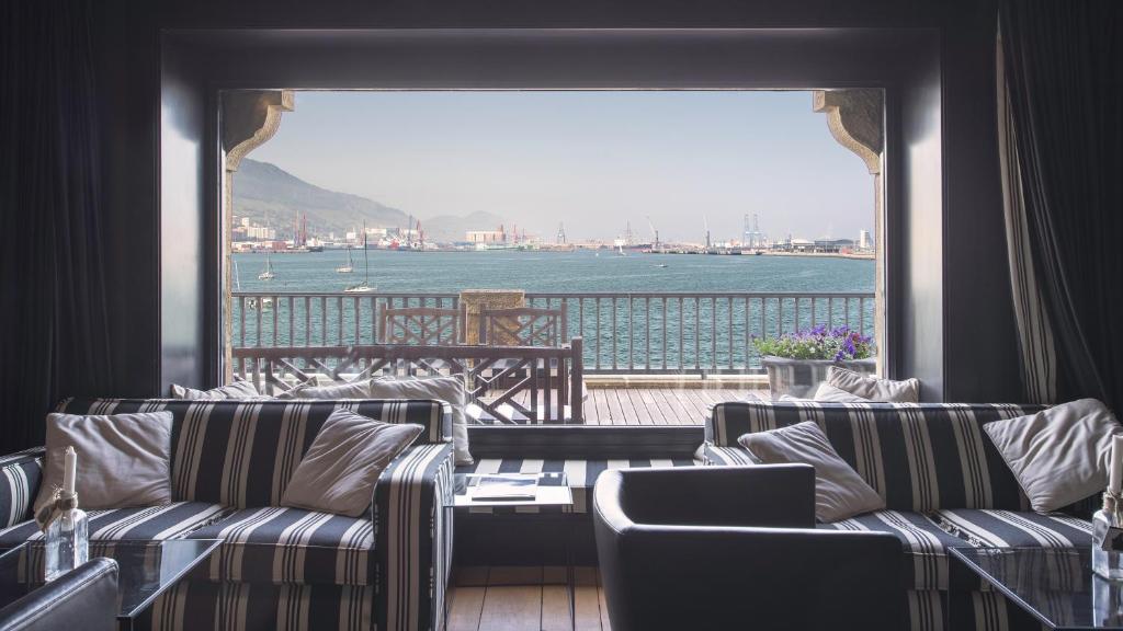 8 Unique Boutique Hotels in Bilbao Spain 31 A cozy lounge in one of the boutique hotels in Bilbao features striped sofas and a coffee table, looking out through a large window to a scenic harbor with boats, distant mountains, and an outdoor deck with wooden chairs and flower pots.