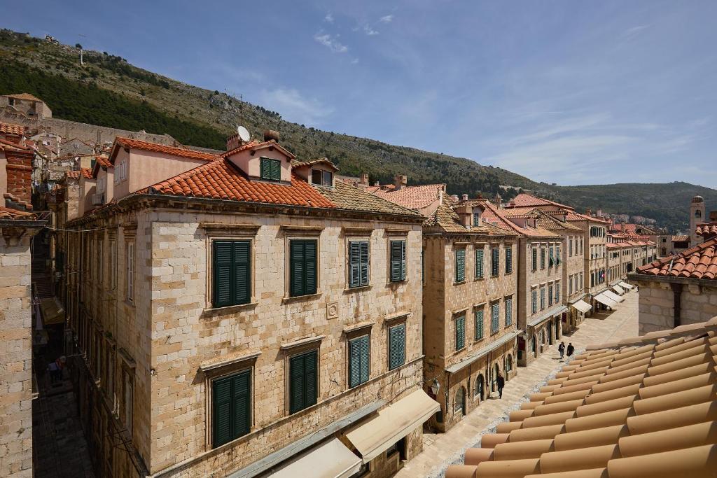 Most Unique Boutique Hotels in Dubrovnik 24 A row of historic stone buildings with red-tiled roofs lines a narrow street in a European town, reminiscent of the charm found at Boutique Hotels in Dubrovnik, with green hills rising in the background under a partly cloudy sky.