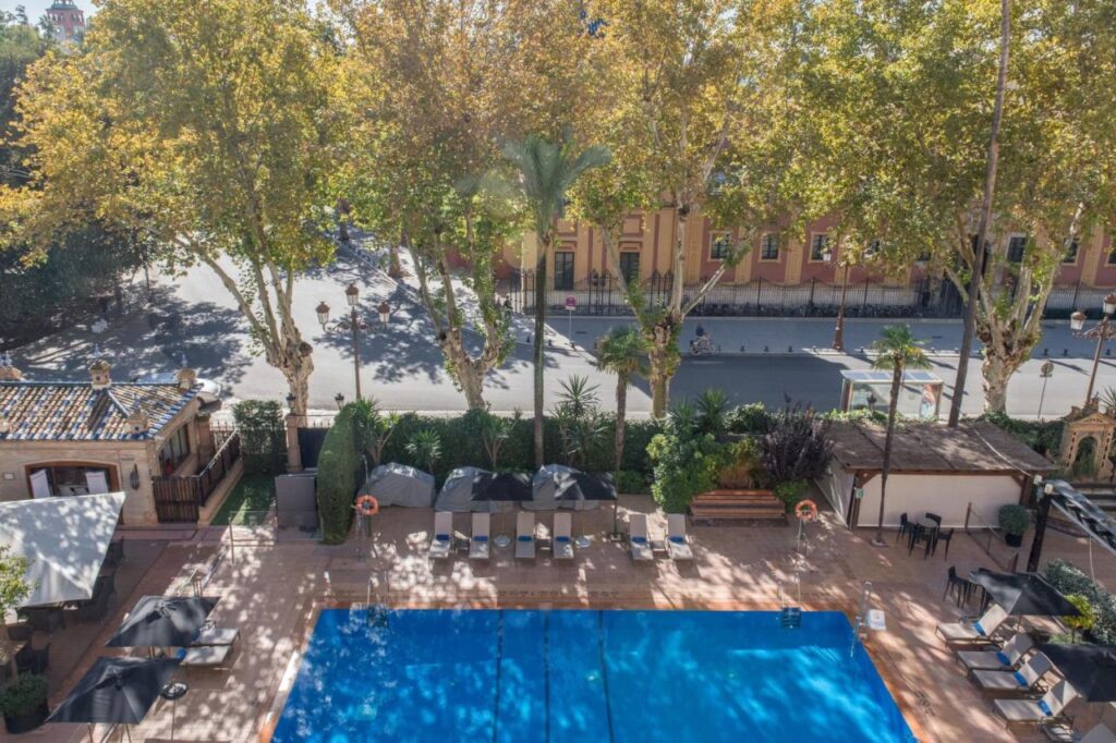 10 UNIQUE Hotels with Pool in Seville 24 A view from above of a rectangular outdoor swimming pool with blue water at one of the top hotels with pool in Seville, surrounded by lounge chairs, umbrellas, and trees. A quiet street and historic buildings are visible in the background.