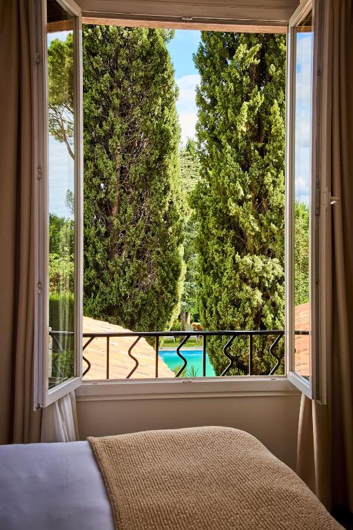 11 Unique Places to Stay in Provence 37 A cozy bed with a tan blanket sits before an open window, revealing tall green trees, a balcony railing, and a glimpse of a blue swimming pool—one of the unique places to stay in Provence under a partly cloudy sky.