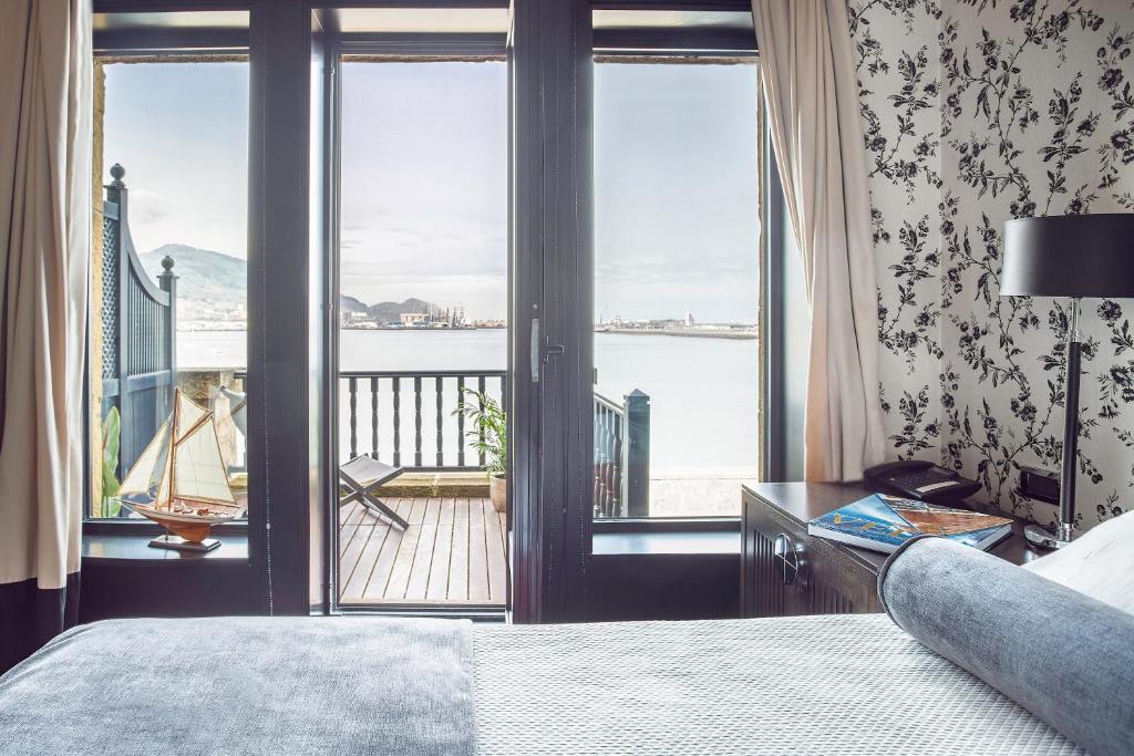 8 Unique Boutique Hotels in Bilbao Spain 33 A bright bedroom in the style of Boutique Hotels in Bilbao, with floral wallpaper and a large bed, features glass doors opening to a balcony overlooking a calm waterfront and distant hills. A small table and chair await on the balcony.