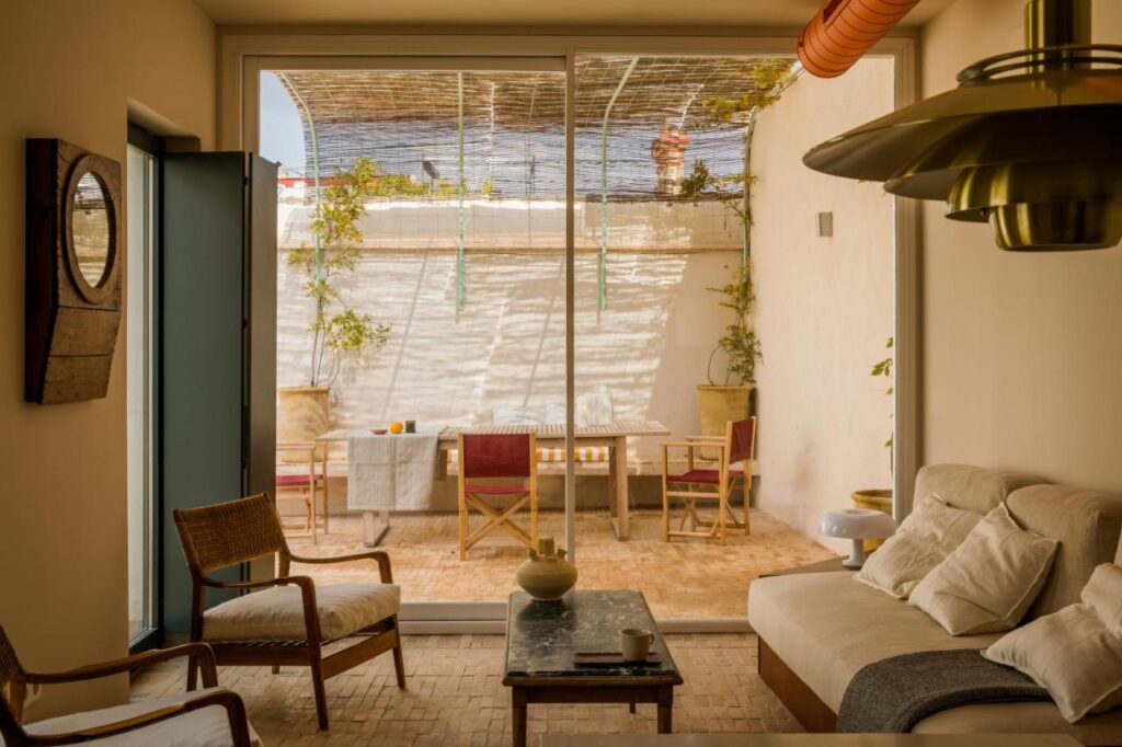 10 UNIQUE Hotels with Pool in Seville 39 Sunlit living room with cozy seating, a wooden chair, and a beige sofa, opening to a patio with a dining table and potted plants. Large glass doors let in natural light—echoing the inviting charm of hotels with pool in Seville.