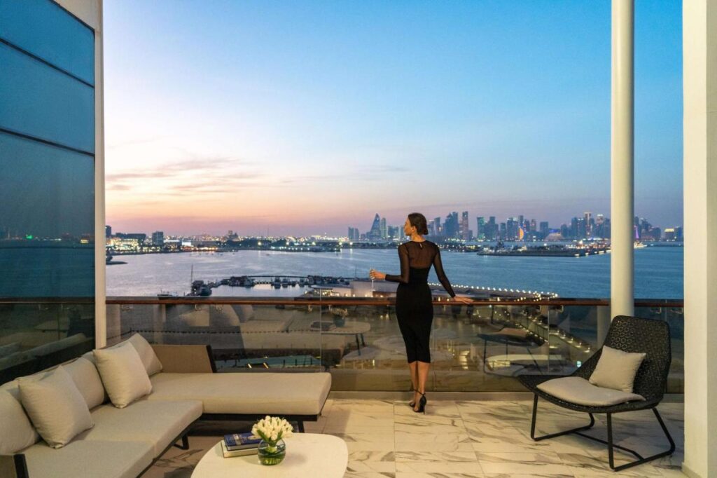 Where to Stay in Doha, Qatar: Transit, City & Beach Hotels 27 A woman in a black dress stands on a modern balcony overlooking the city skyline at sunset—a glimpse into luxurious living and inspiration for those seeking where to stay in Doha, with water views and elegant furnishings all around.