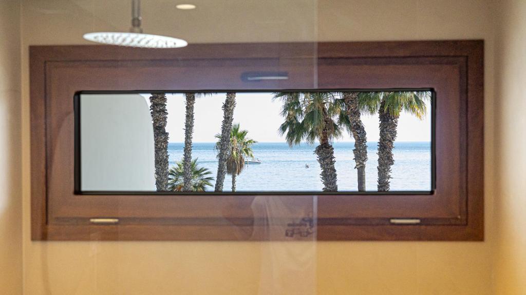 Adults-Only Hotels in Costa del Sol 6 A rectangular wooden-framed window in an adult only hotel in Costa del Sol offers a view of palm trees and the ocean under a clear sky; part of a showerhead and bathroom are visible in the foreground.
