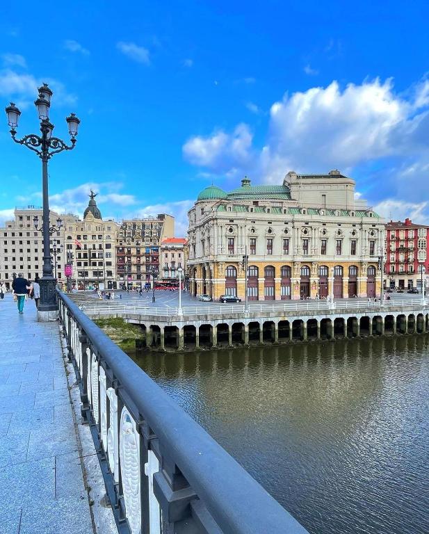 8 Unique Boutique Hotels in Bilbao Spain 13 A riverside view of a historic building with a green roof in a European city, seen from a bridge with ornate lampposts. The sky is blue with scattered clouds, and people stroll along the path near boutique hotels in Bilbao.