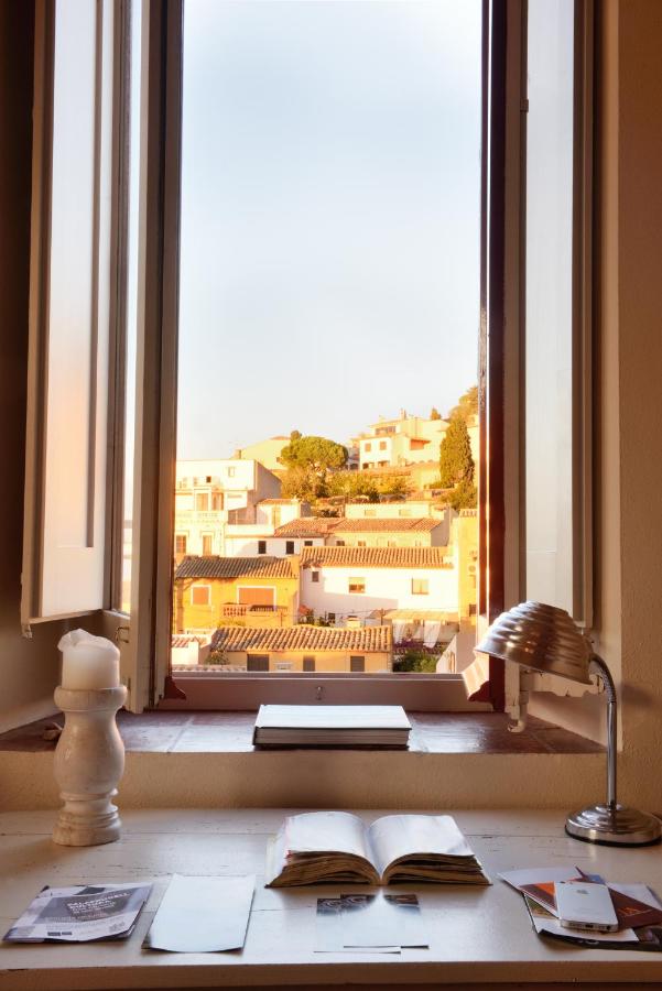 A sunlit desk with an open book, papers, a lamp, and a candle sits by an open window in one of the charming boutique hotels in Costa Brava, overlooking a peaceful town with white and terracotta-roofed buildings under a clear sky.