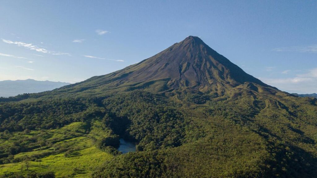Unique Hotels in Costa Rica: Top Stays 5 A large, cone-shaped volcano rises above a lush green forest under a clear blue sky, with shadows and sunlight highlighting the mountain’s slopes and a small pond visible among the trees—an iconic view near unique hotels in Costa Rica.