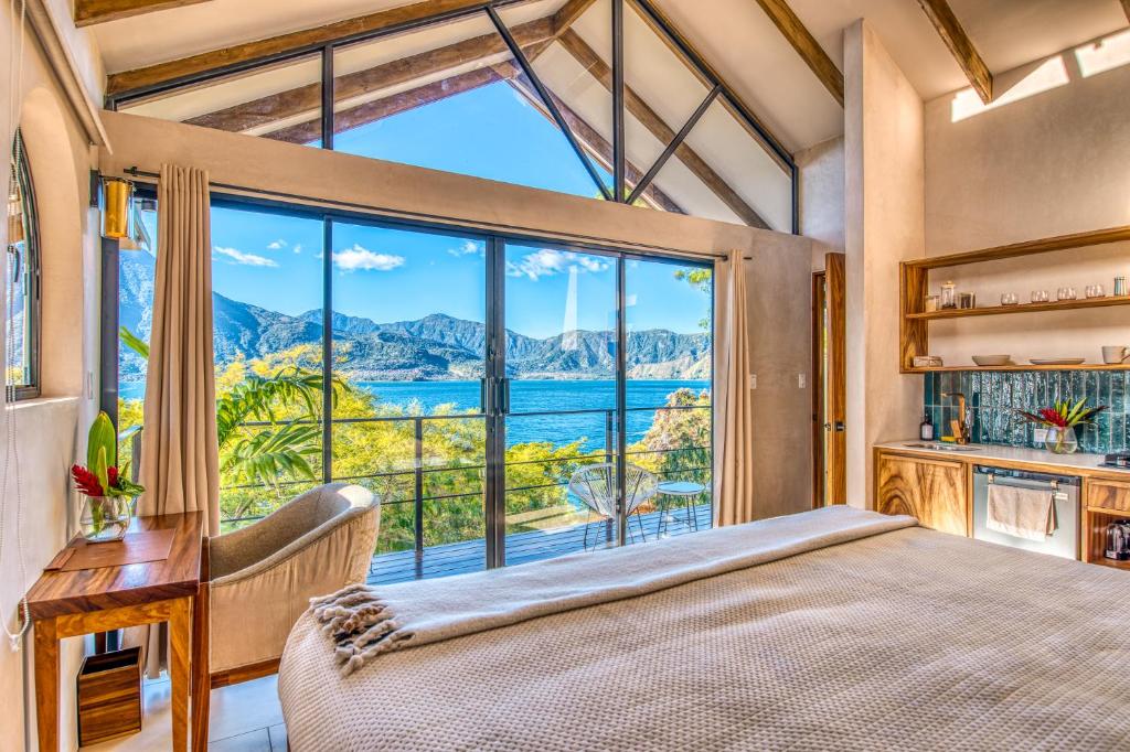 8 UNIQUE Places Where to Stay in Lake Atitlán 27 A cozy bedroom with large glass doors opens to a balcony overlooking a beautiful blue lake and green mountains under a clear sky—an inviting choice for those seeking where to stay in Lake Atitlan. Wooden furniture, plants, and natural light complete the scene.