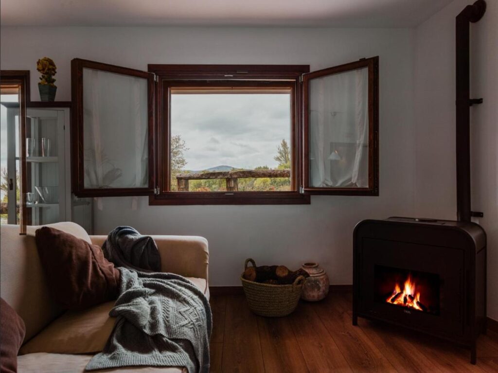 25 Weekend Trips from Madrid Max. 2h Away 76 A cozy living room with a lit fireplace, a beige sofa draped with a gray blanket and brown pillows, a basket of firewood, and an open window revealing the scenic hills—perfect inspiration for weekend trips from Madrid.