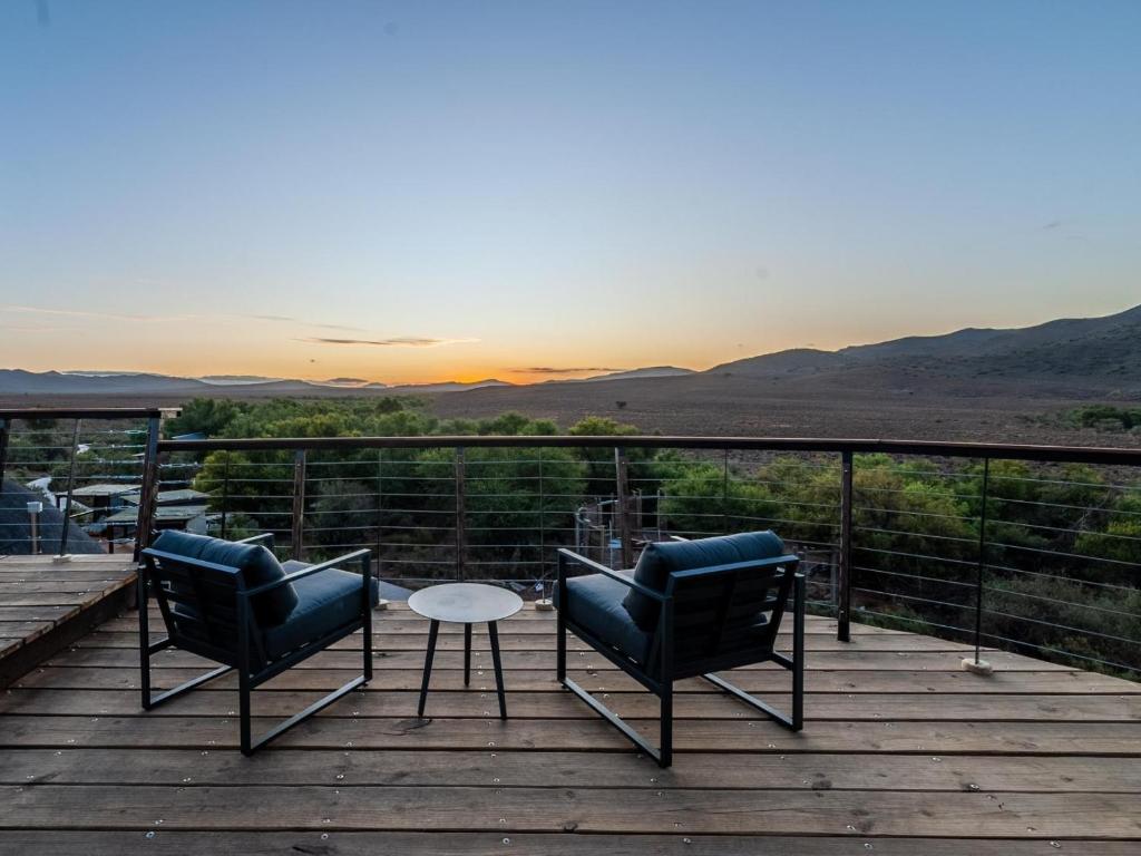 Two black chairs and a small round table sit on a wooden deck at one of the game lodges in Western Cape, overlooking scenic hills and trees at sunset, with mountain silhouettes framing the view under a clear sky.