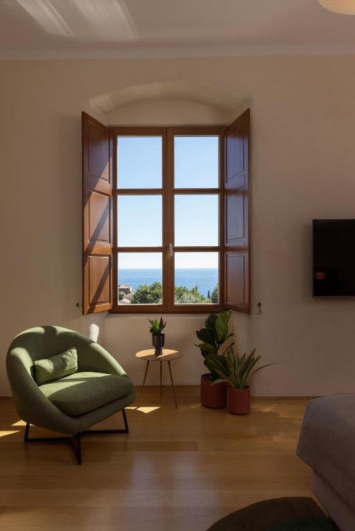 Most Unique Boutique Hotels in Dubrovnik 18 A cozy room in one of the boutique hotels in Dubrovnik features a green armchair, small round table, and potted plants by a large wooden window with open shutters framing the sea and blue sky outside. Sunlight streams onto the wooden floor.