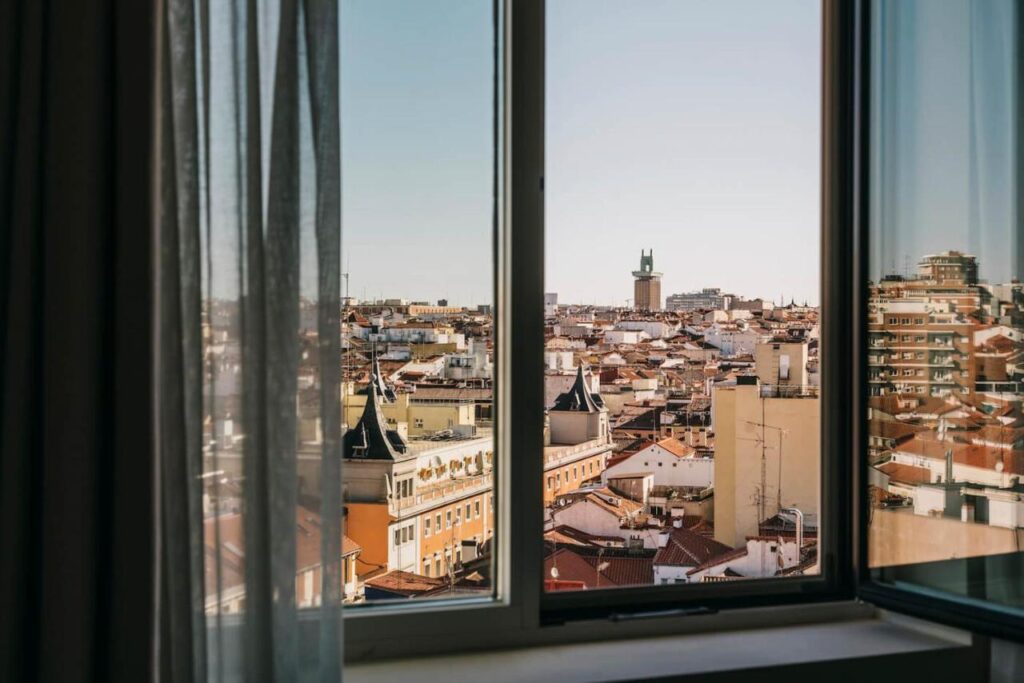 Where to Stay in Madrid: 14 Best Hotels 39 A cityscape view through an open window with sheer curtains on the left side, showing buildings with red-tiled roofs under a clear blue sky—perfect inspiration for those wondering where to stay in Madrid first time visitors.